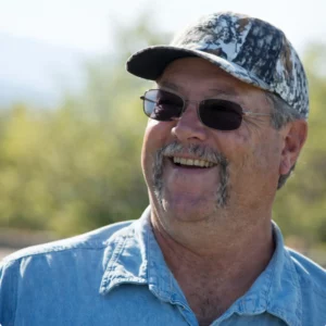 Man with sunglasses and camouflage cap smiling outdoors in a blue denim shirt.