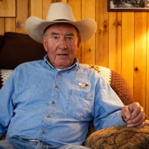 Elderly man wearing a cowboy hat and blue shirt, seated in a wooden-paneled room.