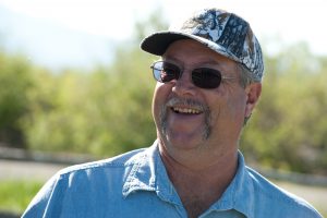 Man wearing sunglasses and camouflage cap, smiling outdoors.
