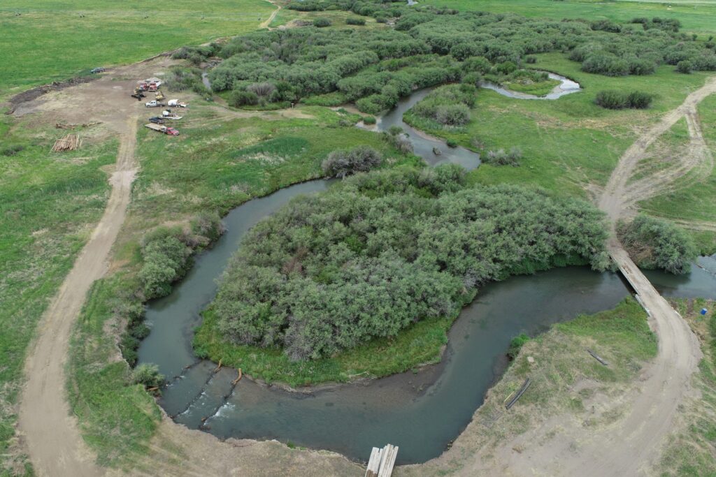 Aerial view of winding river through green fields and trees, with dirt roads and parked vehicles nearby.