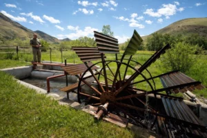 Person standing near a water wheel in a grassy landscape with hills and a blue sky.