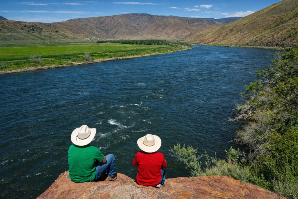 Two people in cowboy hats sit on a rock overlooking a wide river and mountainous landscape.
