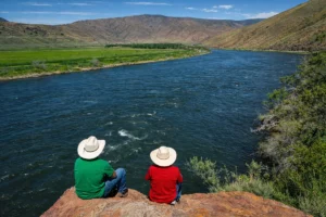 Two people in cowboy hats sit on a rock overlooking a wide river and mountainous landscape.