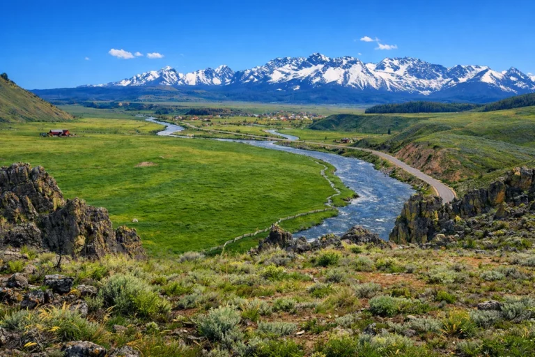 River winding through green valley with rocky hills and snowy mountains under a clear blue sky.